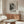 May include: Abstract black and white art poster with Pollock text and exhibition details, displayed above wooden mid-century sideboard in neutral living room. White upholstered chair and glass coffee table visible. Poster features splattered paint texture in monochrome tones with bold typography below the artwork.