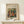 May include: Framed art print of Renoir's impressionist painting showing figures in a outdoor scene with warm colors of orange, green, and blue. The wooden frame sits on a light wood shelf next to a small wrapped white gift box with twine and greenery. Cream colored wall background with soft natural lighting.