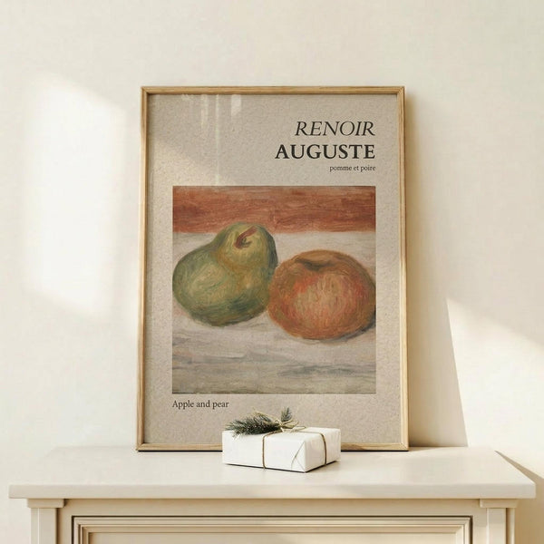 May include: Framed art print featuring Renoir's still life painting of a green pear and red apple on a wooden surface, displayed on a light wood console table next to a small white wrapped gift with evergreen sprig. The poster has beige background with black text reading "RENOIR AUGUSTE" and "Apple and pear" in a natural wood frame.