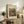 May include: Framed art print of Renoir's impressionist painting showing figures in a pastoral scene with warm earth tones and soft brushstrokes, displayed on rustic wooden sideboard alongside fern plant, ceramic vase, and stacked books in neutral-toned living room setting