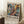 May include: Framed abstract art poster with vibrant blue, orange, yellow and pink paint splatters in Jackson Pollock style, leaning against white wall corner on textured cream rug. Text reads "POLLOCK EMBRACE THE CHAOTIC REALM OF CREATIVITY" with exhibition dates. Coffee cup on wooden block nearby, soft natural lighting creates shadows across the scene.