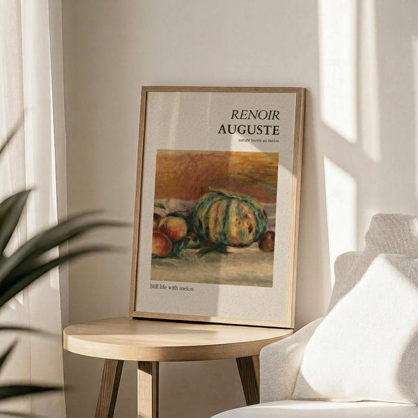 May include: Framed art print featuring Renoir's still life painting of melons and fruit in warm orange and green tones, displayed on a round wooden side table next to a beige upholstered chair. Natural sunlight streams across the neutral-toned room, creating soft shadows on white walls. A plant's green leaves are partially visible in the corner.