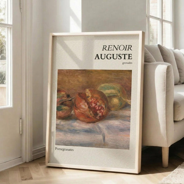 May include: Framed art print leaning against beige upholstered chair in bright living room with white walls and windows. Print shows Renoir's impressionist still life painting of reddish pomegranates on draped fabric, with cream matting and "RENOIR AUGUSTE grenades" text above artwork. Natural wood frame catches window light on hardwood floors.