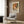 May include: Abstract art poster with vibrant orange, red, green and white paint splatters in Pollock style, displayed in light wooden frame on wood-paneled wall. Modern beige armchair with geometric patterned pillow sits below artwork, next to brass floor lamp and dark round side table with open book.