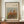 May include: Framed art print displaying an impressionist-style painting of autumn trees with orange, red, and green foliage against a pale blue sky. The artwork features warm earth tones and loose brushstrokes typical of impressionist technique. Shown in a thin brown wooden frame with cream matting, displayed on a wooden sideboard next to a white ceramic vase and stacked books in an elegant room with white wainscoting and cream walls.