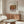 May include: Renoir art poster displaying painted pomegranates in warm reds and greens, framed and hung above a wooden sideboard in a minimalist living room with cream walls, white upholstered chair, glass coffee table, and modern brass floor lamp