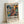 May include: Framed abstract art poster with vibrant swirling paint strokes in teal, coral, yellow and black, featuring exhibition-style text reading "POLLOCK" and "EMBRACE THE CHAOTIC REALM OF CREATIVITY" with May-August 2030 dates. Light wood frame leaning against white wall on light surface, showing texture of paper print and natural lighting creating soft shadows.