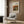 May include: Beige armchair with geometric patterned pillow positioned next to gold floor lamp in modern living room. Framed art poster showing Renoir still life painting with melon and fruits hangs on wood-paneled wall. Natural lighting from window with sheer curtains, dark round side table with open book visible.