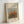 May include: Framed art print showing an impressionist painting of autumn trees with orange, red, and green foliage against a pale blue sky. The artwork is displayed in a light wood frame with white matting, featuring text identifying it as a Pierre Auguste Renoir painting titled "grove of trees." The frame leans against a white wall on a light surface.