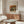 May include: Framed Renoir still life poster featuring green striped melon and fruits in warm earth tones, displayed above walnut sideboard in minimalist living room with cream chair, glass coffee table, and white table lamp. Soft natural lighting creates gentle shadows on pale walls.