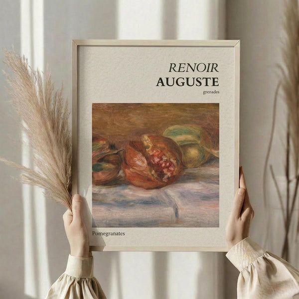 May include: Hands holding a framed art print of Renoir's pomegranate still life painting showing split red pomegranates with exposed seeds on a soft fabric surface. The poster has cream matting with "RENOIR AUGUSTE grenades" text above and "Pomegranates" below the artwork. Natural lighting creates soft shadows on white curtains in background, with dried pampas grass visible to the left.