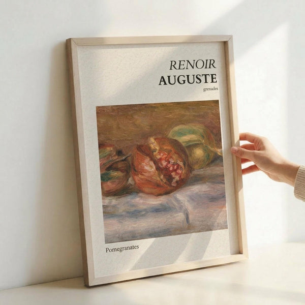May include: Framed art print featuring Renoir's impressionist painting of split pomegranates with ruby red seeds visible, accompanied by green fruits on draped fabric. The artwork shows warm earth tones and soft brushstrokes typical of the period. Print has white matting with "RENOIR AUGUSTE" and "grenades" text above the image, "Pomegranates" below. Natural wood frame held by hand against white wall, showing realistic room display mockup.