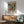 May include: Abstract colorful artwork poster in wood frame hanging above modern wooden sideboard with white table lamp. Mint green mid-century armchair with wooden arms sits nearby with small round coffee table. Bright natural light creates shadows on white wall. Poster features vibrant blues, oranges, yellows and reds in chaotic painterly style with text below the artwork.
