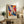 May include: Framed abstract art print with vibrant swirling colors in blues, oranges, reds, and yellows displayed on rustic wooden sideboard. Scene shows cream chair with brown throw pillow, potted fern, stacked books, ceramic vase, and neutral wall background creating cozy living room setting.