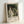 May include: A framed exhibition poster featuring Renoir's "La Promenade" from 1870, showing a woman in a flowing white dress and a man in dark clothing walking through a lush green landscape. The poster has white text reading "PIERRE AUGUSTE exhibition" below the impressionist painting. The light wood frame leans against a white wall, casting soft shadows in natural lighting.