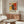 May include: Abstract expressionist-style poster with vibrant red, yellow, and black paint splatters and geometric lines, displayed in thin wooden frame above mid-century walnut credenza. White sculptural chair and glass coffee table in foreground reflect the artwork. Minimalist room setting with beige walls and natural lighting creates warm, contemporary gallery atmosphere.