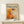 May include: Framed abstract art poster featuring bold orange and white paint splatters with black accents in Jackson Pollock's signature drip style, displayed on light wood console table next to small wrapped gift with evergreen sprig against cream wall