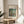 May include: Framed art print featuring Pierre Auguste Renoir's impressionist painting with lush green foliage, figures near water, and warm earth tones. Displayed on light wooden round side table next to beige upholstered chair with decorative pillow. Natural sunlight streams across neutral wall, casting soft shadows from nearby plant leaves.
