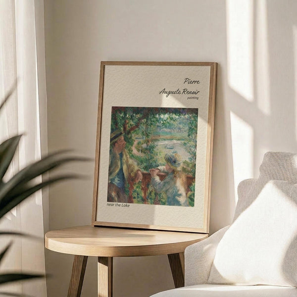 May include: Framed art print featuring Pierre Auguste Renoir's impressionist painting with lush green foliage, figures near water, and warm earth tones. Displayed on light wooden round side table next to beige upholstered chair with decorative pillow. Natural sunlight streams across neutral wall, casting soft shadows from nearby plant leaves.