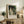 May include: Framed art print featuring a couple in a lush green garden setting, with the woman in a flowing white dress and man in dark clothing. The impressionist-style painting shows rich greens and earth tones. The poster displays "PIERRE AUGUSTE exhibition" text below the artwork with "1870" and "La Promenade" details. Styled on a rustic wooden sideboard with books, ceramic vase, fern plant, and cream upholstered chair visible in a neutral-toned living space.