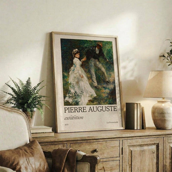 May include: Framed art print featuring a couple in a lush green garden setting, with the woman in a flowing white dress and man in dark clothing. The impressionist-style painting shows rich greens and earth tones. The poster displays "PIERRE AUGUSTE exhibition" text below the artwork with "1870" and "La Promenade" details. Styled on a rustic wooden sideboard with books, ceramic vase, fern plant, and cream upholstered chair visible in a neutral-toned living space.