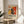 May include: Abstract expressionist art poster in wooden frame displayed on round wooden side table next to beige upholstered chair. Poster features bold splashes of red, yellow, black and white paint with dynamic brushstrokes and splatters. Exhibition-style design with "POLLOCK" text and dates "MAY 18, 2030 - AUGUST 14, 2030" below colorful abstract artwork. Soft natural lighting creates shadows on cream wall behind, with green plant leaves visible at left edge.