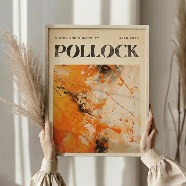 May include: Person in cream blouse holding framed abstract art poster featuring orange, white, and black paint splatters in Jackson Pollock's drip painting style. Poster displays "CHAOS AND CREATIVITY" and "1912-1956" text above "POLLOCK" in bold letters. Light wood frame against white curtained background with dried pampas grass visible on left side.
