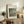 May include: Framed Renoir reproduction print showing figures by a lake in impressionist style with green foliage and soft brushstrokes, displayed on rustic wooden sideboard alongside fern plant, ceramic vase, and stacked books in neutral-toned living room setting with cream upholstered chair visible.