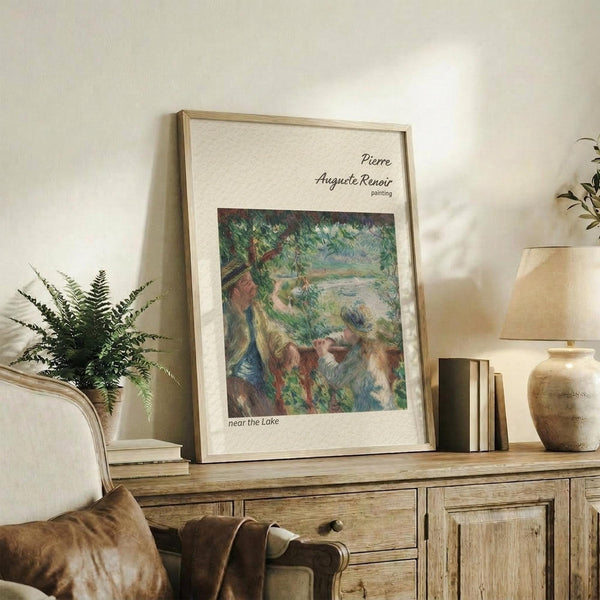 May include: Framed Renoir reproduction print showing figures by a lake in impressionist style with green foliage and soft brushstrokes, displayed on rustic wooden sideboard alongside fern plant, ceramic vase, and stacked books in neutral-toned living room setting with cream upholstered chair visible.