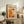 May include: Framed abstract art poster with orange, white and black paint splatters displayed on rustic wooden sideboard alongside potted fern, ceramic vase, stacked books, and leather armchair in neutral-toned living room setting