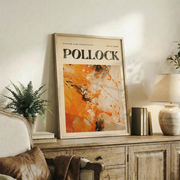 May include: Framed abstract art poster with orange, white and black paint splatters displayed on rustic wooden sideboard alongside potted fern, ceramic vase, stacked books, and leather armchair in neutral-toned living room setting