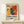 May include: Framed abstract art poster in light wood frame leaning against white wall on natural wood shelf. Features vibrant splattered paint design with red, yellow, black and white colors in Jackson Pollock style. Text reads "POLLOCK" and "EMBRACE THE CHAOTIC REALM OF CREATIVITY" with exhibition dates. Small white wrapped gift box with twine and pine sprig sits beside frame.