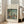 May include: Framed poster print leaning against white wall next to beige armchair in bright living room. Print shows Impressionist-style painting of figures by lakeside with lush green foliage, rendered in soft blues, greens, and earth tones. Light wood frame with cream matting. Natural sunlight streams through windows onto hardwood floors.