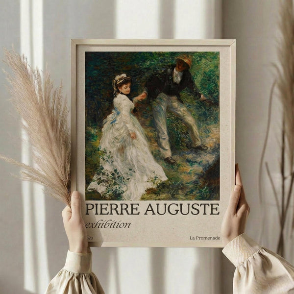 May include: Hands holding a framed reproduction poster featuring an Impressionist painting of a woman in white dress and man in dark clothing sitting together in dappled forest light. The beige-framed print shows "PIERRE AUGUSTE exhibition" and "La Promenade" text below the artwork. Person wearing cream ruffled sleeves holds the poster against white curtained window, with dried pampas grass visible to the left.