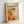 May include: Framed abstract art print leaning against white wall corner, featuring bold orange and black paint splatters in Jackson Pollock's drip painting style. Light wood frame contains cream-colored poster with "POLLOCK" in large black letters, "CHAOS AND CREATIVITY 1912-1956" subtitle, and dynamic abstract artwork below with energetic brushstrokes and paint drips in warm orange, black, and white tones.