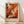 May include: Framed abstract expressionist poster with vibrant orange, red, and black paint splatters and drips on white background, displayed against white textured wall on light wood flooring with natural sunlight creating diagonal shadows across the scene.