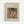 May include: Framed art print displaying Pierre-Auguste Renoir's painting of a woman in a pink dress and dark feathered hat, seated at a table with a blue and white teacup. The reproduction shows warm, soft brushstrokes typical of Impressionist style. Light wood frame with cream matting displays text identifying the artist and artwork title. Wall-mounted against neutral background.