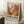 May include: Framed abstract art exhibition poster with swirling paint in warm oranges, reds, and yellows against cream background, leaning against white wall on textured cream rug. Coffee cup on wooden block beside poster. Natural window light creates soft shadows across the cozy interior scene.
