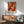 May include: Abstract expressionist artwork with vibrant red, orange, and black paint splatters and drips on white background, displayed in thin wooden frame above mid-century modern wooden credenza. Scene features sage green upholstered armchair with wooden arms, white ceramic table lamp, and circular wooden side table. Warm natural sunlight streams across white wall creating geometric shadows.