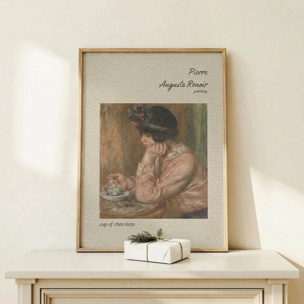 May include: A framed art print featuring a woman in pink clothing and dark hat drinking from a teacup, displayed on a light wood surface beside a small wrapped gift with greenery. The artwork shows muted earth tones and impressionist painting style, housed in a thin wooden frame with cream matting and text above and below the image.
