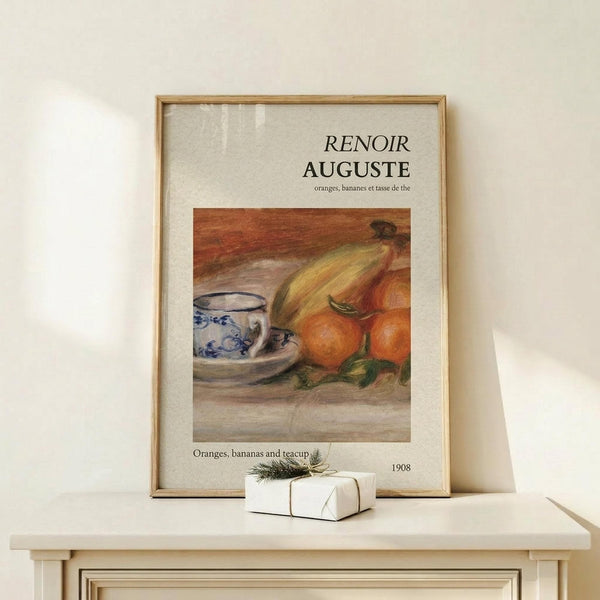 May include: Framed art print displaying Renoir's still life painting of oranges, bananas, and blue-and-white patterned teacup on wooden surface. Warm orange and yellow tones dominate the artwork. Natural wood frame sits on light wooden shelf beside small white wrapped gift with twine and greenery sprig. Cream colored wall background with soft natural lighting.