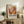 May include: Framed abstract art exhibition poster with warm orange, yellow and red paint splashes displayed on wooden sideboard beside fern plant, books, and ceramic vase in cozy living room setting with beige armchair