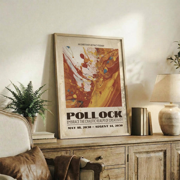 May include: Framed abstract art exhibition poster with warm orange, yellow and red paint splashes displayed on wooden sideboard beside fern plant, books, and ceramic vase in cozy living room setting with beige armchair