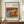 May include: Framed vintage-style art print featuring Renoir's still life painting of oranges, bananas, and a blue and white patterned teacup displayed on a wooden sideboard. The warm-toned artwork shows soft brushstrokes in yellows, oranges, and blues against a cream background. The print has a thin brown frame and sits beside a small white ceramic vase and stacked books in an elegant room with white paneled walls.
