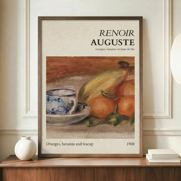 May include: Framed vintage-style art print featuring Renoir's still life painting of oranges, bananas, and a blue and white patterned teacup displayed on a wooden sideboard. The warm-toned artwork shows soft brushstrokes in yellows, oranges, and blues against a cream background. The print has a thin brown frame and sits beside a small white ceramic vase and stacked books in an elegant room with white paneled walls.