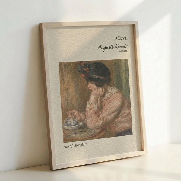 May include: Framed art print displayed on white surface against white wall. Light wood frame contains reproduction of impressionist painting showing woman in pink dress and dark feathered hat, resting chin on hand while holding delicate teacup. Soft, muted colors in classical French painting style. Text above reads "Pierre Auguste Renoir painting" and below "cup of chocolate" on cream textured background.