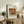 May include: Framed art print displaying a still life painting of oranges, bananas, and a blue and white patterned teacup on a wooden surface. The poster features "RENOIR AUGUSTE" text at top with "Oranges, bananas and teacup" and "1908" below the artwork. Shown in thin wooden frame, leaning against white wall on rustic wooden sideboard alongside fern plant, books, and ceramic vase in neutral-toned living room setting.