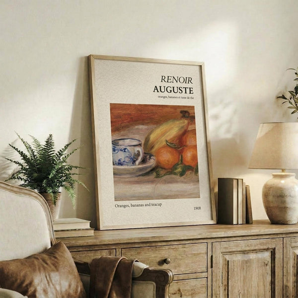 May include: Framed art print displaying a still life painting of oranges, bananas, and a blue and white patterned teacup on a wooden surface. The poster features "RENOIR AUGUSTE" text at top with "Oranges, bananas and teacup" and "1908" below the artwork. Shown in thin wooden frame, leaning against white wall on rustic wooden sideboard alongside fern plant, books, and ceramic vase in neutral-toned living room setting.