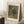 May include: Framed art print leaning against white wall corner showing floral still life painting with wildflowers in earthenware vase. Print has cream matting with black text identifying artist and date. Scene includes wooden side table with white coffee cup, beige throw blanket, and white textured rug. Natural lighting creates soft shadows on wall.