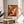 May include: Abstract expressionist artwork poster with vibrant orange, red, white and black paint splatters and brushstrokes, displayed in light wood frame on round wooden side table next to beige upholstered chair. Natural sunlight creates soft shadows on white wall behind. Green plant leaves visible in corner of minimalist, modern living room setting.